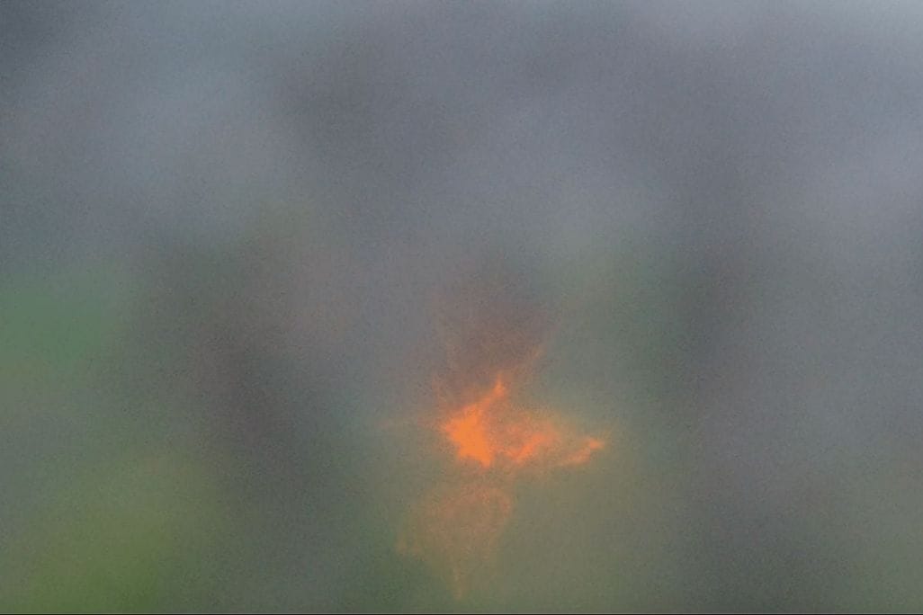 A photograph depicting a soft, misty background with an abstract, glowing orange and yellow light at the center. The light has a starburst pattern, contrasting with the diffuse, muted colors surrounding it.