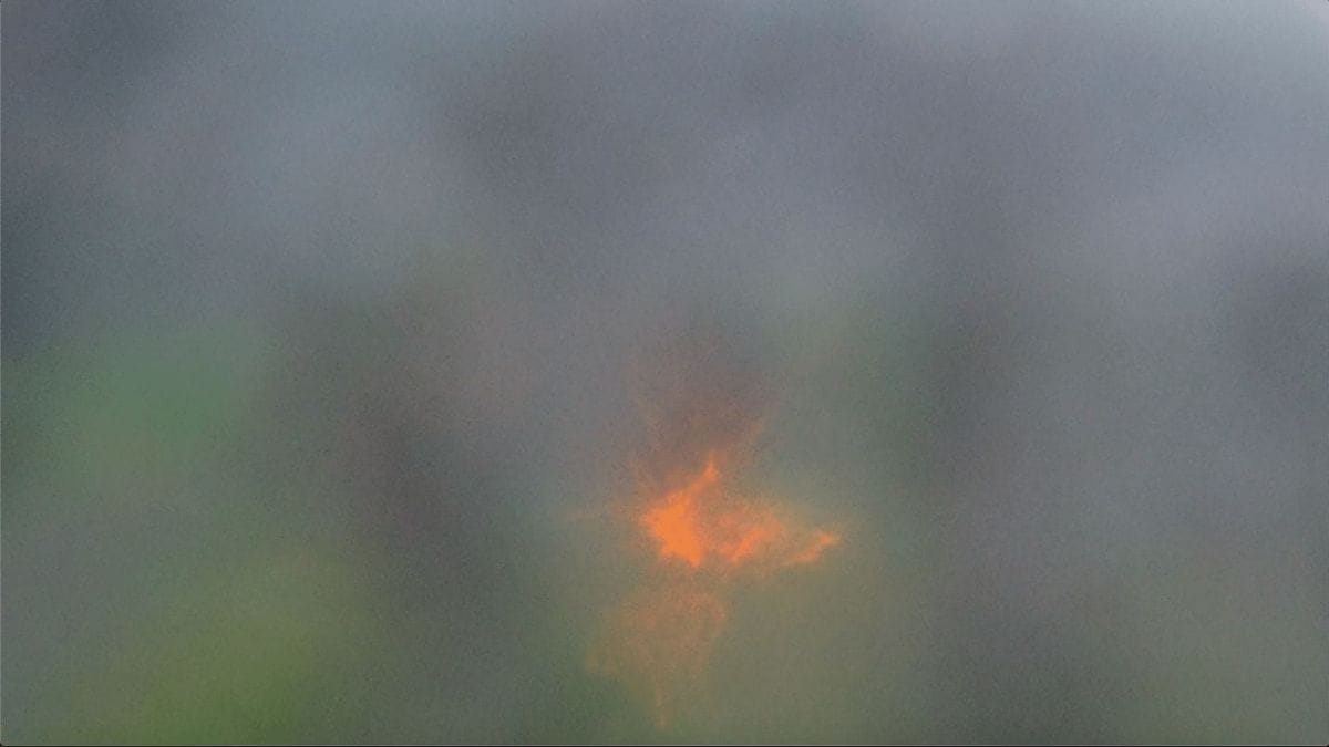 A photograph depicting a soft, misty background with an abstract, glowing orange and yellow light at the center. The light has a starburst pattern, contrasting with the diffuse, muted colors surrounding it.
