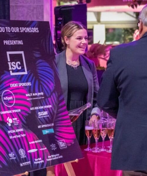 A woman stands and smiles next to a table with champagne glasses, engaging in conversation with a man in a suit at a formal event. Behind them, a large sign thanks the event sponsors, listing various company logos and names. The setting has a festive atmosphere.