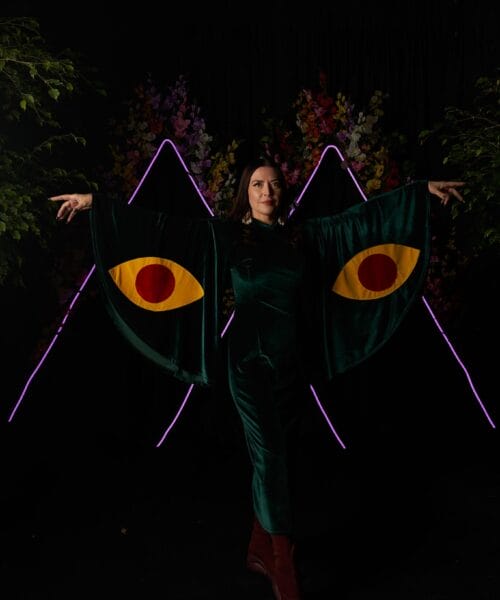 A person wearing a dark green outfit with wing-like sleeves featuring large yellow and red eye designs. They are posing with both arms extended, and a colorful floral arrangement and purple neon lines are visible in the dark background.