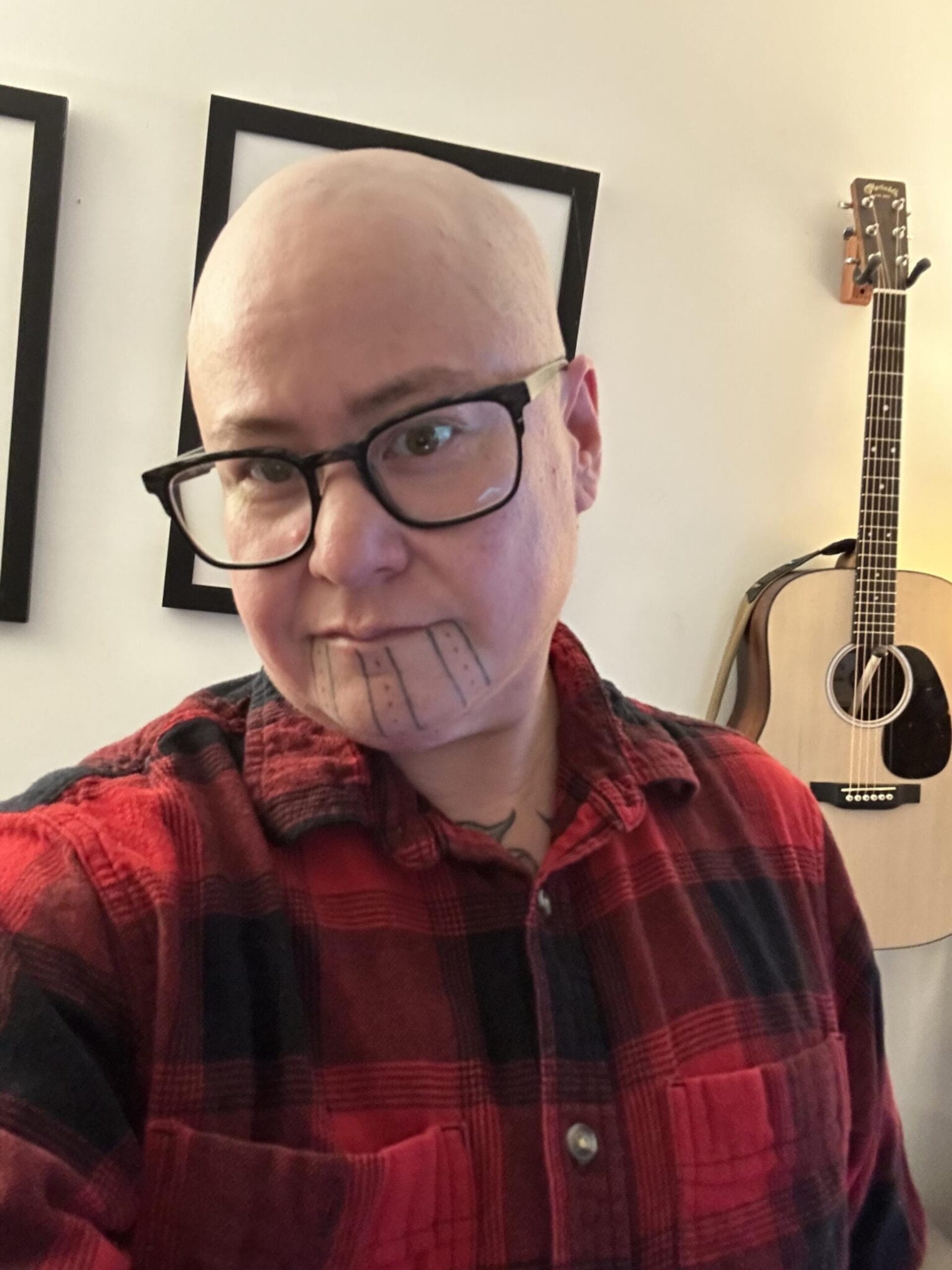 A person wearing glasses and a red plaid shirt stands indoors. They have a bald head and a chin tattoo with lines and shapes. A guitar hangs on the wall in the background.