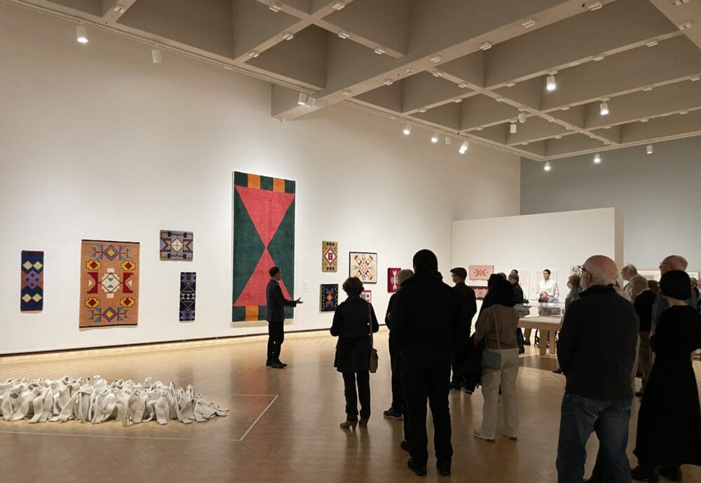 A group of visitors standing in front of large rugs displayed on the wall as a curator describes the work to them.