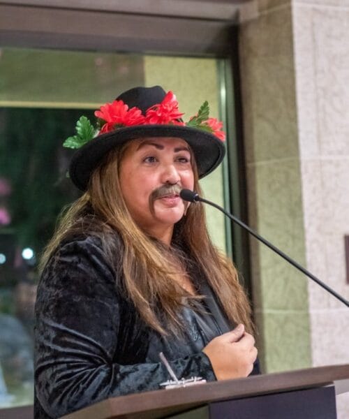 A person in a black jacket and hat, adorned with red flowers and green leaves, stands at a lectern speaking into a microphone. They have long hair and are wearing a fake mustache. The background includes a large window with evening outdoor lighting.