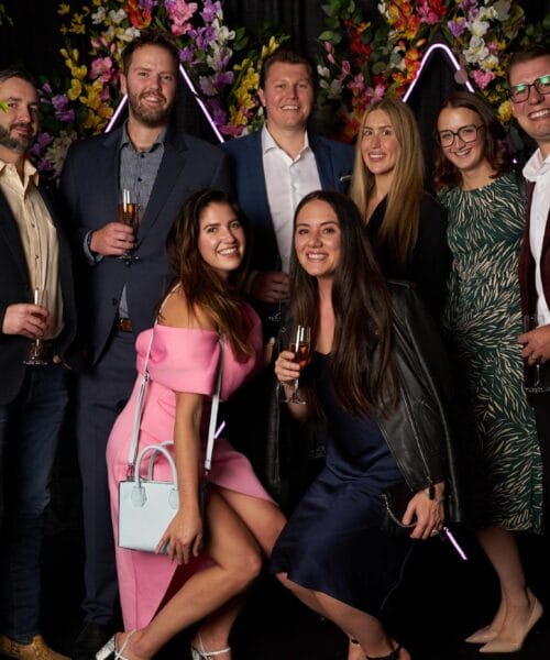 A group of eight people are posing together at an event, smiling at the camera. Five men and three women are dressed in formal attire, holding drinks. There are decorative plants and neon lights in the background. Two women are kneeling in front, smiling brightly.