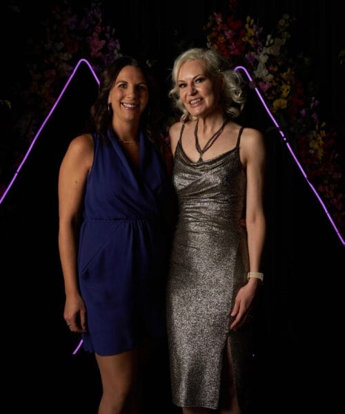 Two women are posing and smiling together in front of a dark backdrop with glowing purple geometric shapes. One woman is wearing a blue sleeveless dress, while the other is in a silver sleeveless dress. Green foliage is visible on both sides of the backdrop.