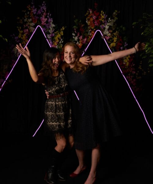 Two women are posing enthusiastically in front of a black backdrop adorned with pink neon mountain outlines and colorful floral arrangements. The woman on the left wears a lace dress and black boots, while the woman on the right wears a dark dress and red shoes.