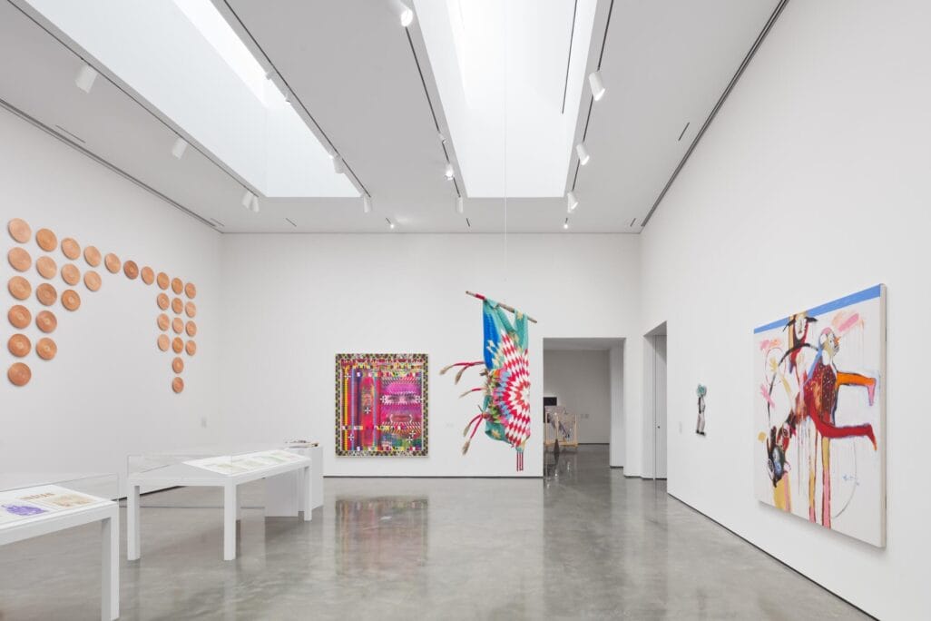 A contemporary art gallery with white walls displays colorful abstract paintings, a feathered artwork hanging, ceramic pieces arranged in a pattern, and art on tables. Enjoy the vibrant scene on RAWLCO Free Day, as polished floors reflect ceiling lights.