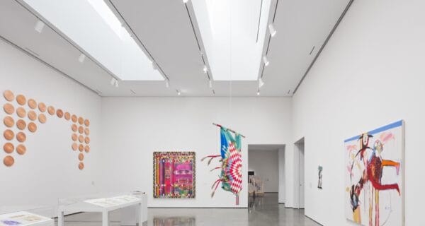 A contemporary art gallery with white walls displays colorful abstract paintings, a feathered artwork hanging, ceramic pieces arranged in a pattern, and art on tables. Enjoy the vibrant scene on RAWLCO Free Day, as polished floors reflect ceiling lights.