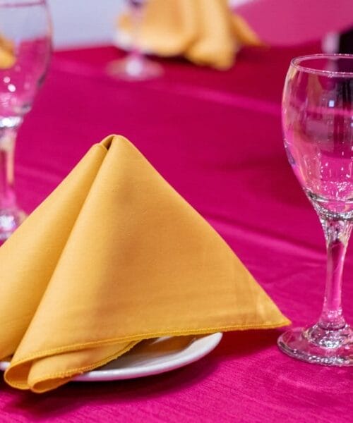 A table setting with a bright pink tablecloth, featuring a neatly folded yellow napkin on a white plate and an empty wine glass. Other similarly set places can be seen in the background, creating a vibrant and elegant dining atmosphere.