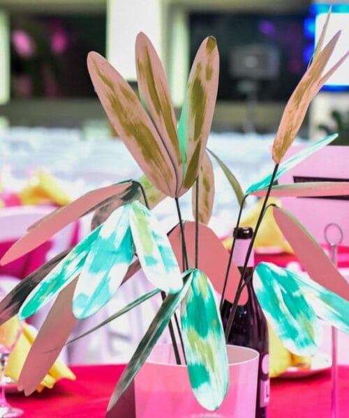 A colorful event table setup featuring a centerpiece with abstract, multicolored decorative leaves. The table is adorned with white chairs, pink tablecloths, yellow napkins, and several wine glasses. The background is softly lit with purple and blue hues.