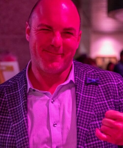 A man in a checkered blazer and light-colored shirt smiles and gives a thumbs-up gesture. The image is illuminated with a pinkish light. The background is out of focus, with other people and event decor visible.