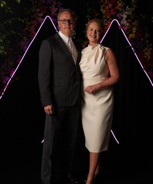 A man and a woman stand smiling in formal attire against a dark backdrop decorated with colorful flowers and illuminated purple neon lines in a triangular shape. The man wears a dark suit and white shirt, while the woman is dressed in a white, sleeveless dress.