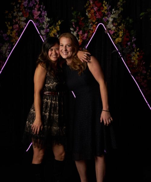 Two women stand side by side with their arms around each other, smiling at the camera. They are dressed in dark-colored dresses, and the backdrop features decorative triangular neon lights and vibrant flowers, with green foliage on either side.