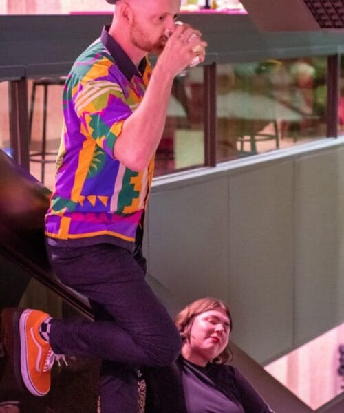 A person in a colorful shirt, black hat, and orange sneakers sits casually on an escalator while drinking from a glass. Another person in a black outfit with white stripes is sitting below on the escalator steps, looking tired or resting.