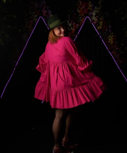 A woman in a bright pink dress and green hat stands with her back to the camera, looking over her shoulder. The background features a dark setting with neon purple triangular lights and floral decorations, creating a vibrant, festive atmosphere.