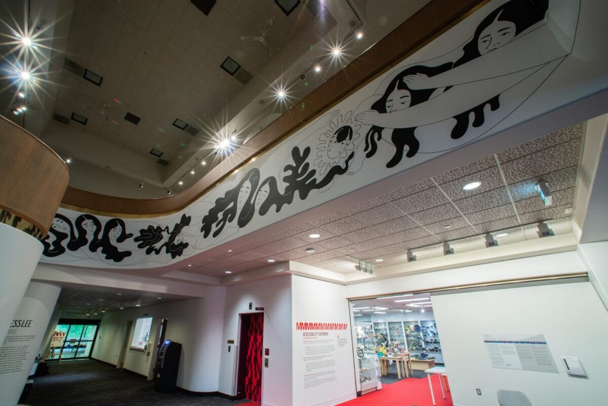 Interior of a modern building with a curved balcony featuring abstract black and white artwork depicting people and organic shapes. Below, there are wall displays, a red carpeted area, and overhead lighting. Displays of various objects are visible.