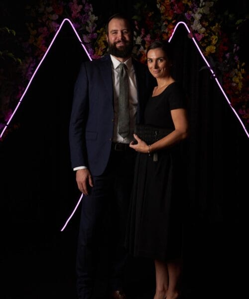 A man in a navy suit with a tie and a woman in a black dress holding a black clutch stand together in front of a dark background with pink neon lights and colorful flowers.