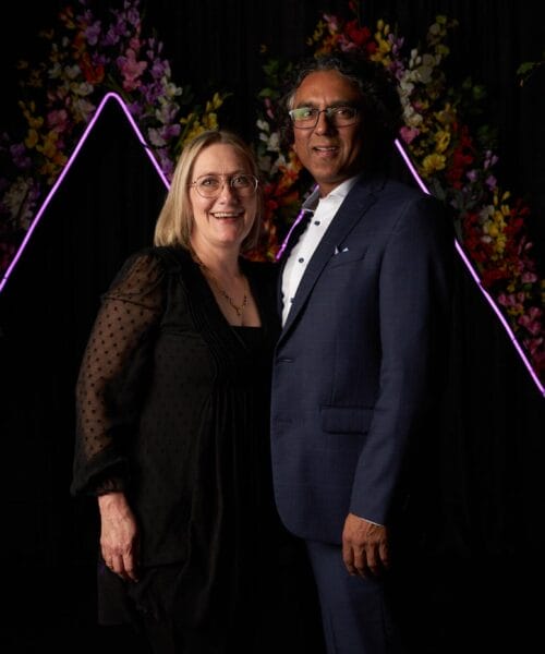 A smiling woman and man stand closely together against a dark backdrop adorned with colorful floral arrangements and illuminated geometric shapes. The woman wears glasses and a black dress, while the man wears glasses and a dark blue suit.