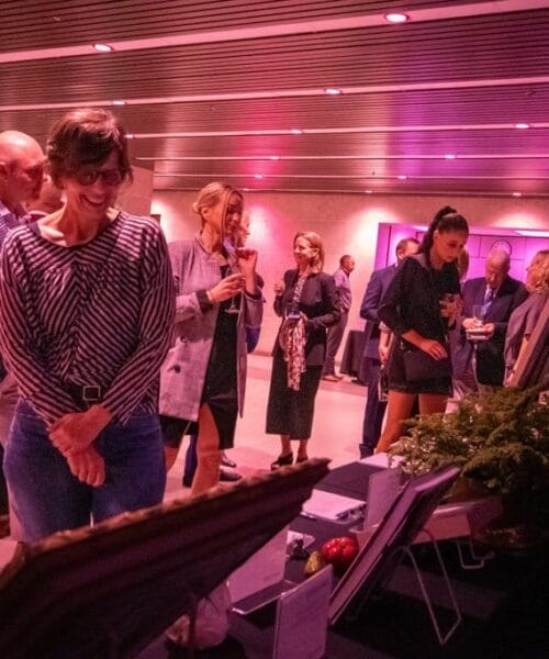 A group of people in semi-formal attire are gathered indoors, engaging with various displays at a well-lit event with a purple ambiance. Some people are looking at framed items on tables, while others chat and sip drinks. Plants and decor adorn the space.
