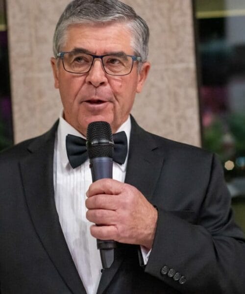 A man with gray hair, wearing glasses, a black tuxedo, and a black bow tie, holds a microphone while speaking. The background features a large window with blurred outdoor scenery.