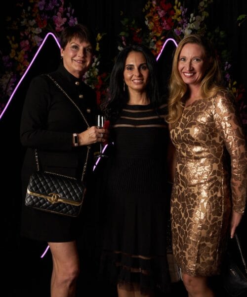 Three women stand together in front of a decorative backdrop with neon pink triangular lights. The woman on the left wears a black dress and holds a black purse, the middle woman wears a black dress, and the woman on the right wears a shiny, gold dress.
