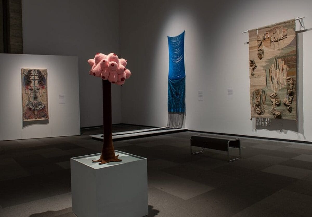Three large textile works are hung on the wall in the background, a tree like sculpture is placed in the middle with crochet and yarn boobs on top.