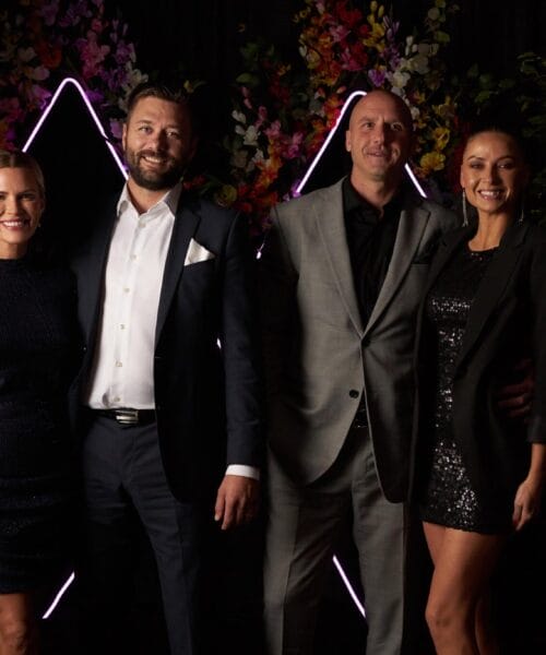 Four people, two women and two men, stand together against a backdrop featuring illuminated geometric shapes and floral decorations. All are dressed in formal attire. The women wear dark, sparkly dresses, and the men are in suits, smiling for the camera.