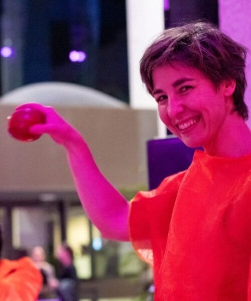 A person with short hair, wearing a bright orange outfit, smiles and holds a red object. The background is lit with pink and purple lights, suggesting an indoor event or performance. Another person in a similar outfit is seen in the background.