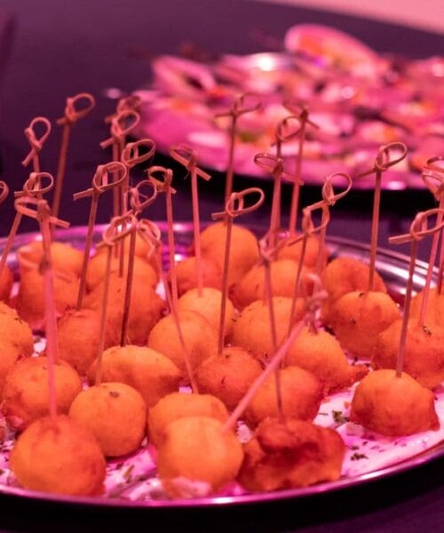 A close-up of a silver platter containing golden-brown fried appetizers, each skewered with toothpicks, on a dark tablecloth. In the background, another platter with bite-sized hors d'oeuvres is slightly out of focus. Stacks of small white plates are placed beside them.