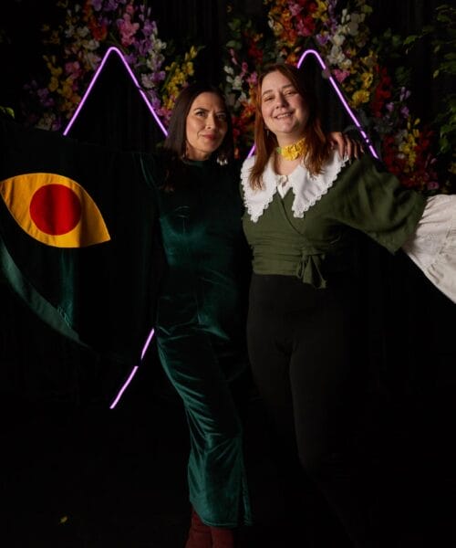 Two women posing together amidst a backdrop of foliage and vibrant lights. The woman on the left is wearing a green dress with a cape featuring a red and yellow eye design. The woman on the right is in a green and white outfit, both smiling and gesturing outward.