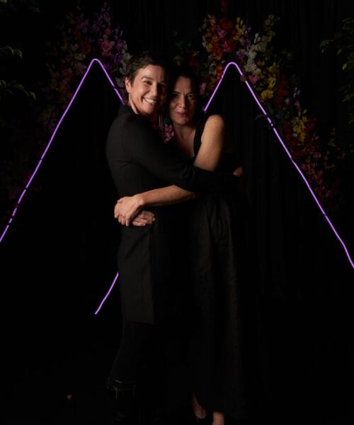 Two people wearing black outfits stand close together in front of a dark backdrop adorned with neon pink triangular lights and colorful flowers. They are smiling and embracing each other, creating a warm and affectionate scene.