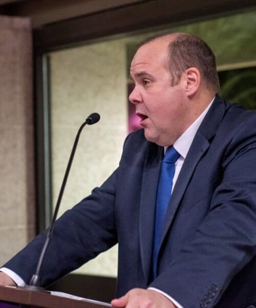 A person in a blue suit and tie is speaking passionately at a podium. The background features a stone wall and large windows, suggesting an indoor setting, such as a conference or event. The person appears engaged in delivering a speech or presentation.