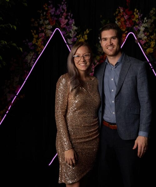 A woman in a sparkly gold dress and glasses stands next to a man in a blue blazer and checked shirt. They both smile and pose in front of a black backdrop adorned with illuminated triangular shapes and colorful flowers. Greenery is visible on both sides.