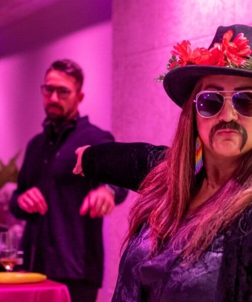 A person wearing a black hat with flowers, sunglasses, and a fake mustache points ahead while standing in a room with purple lighting. In the background, another person is at a DJ booth with a laptop. There are colorful decorations around.