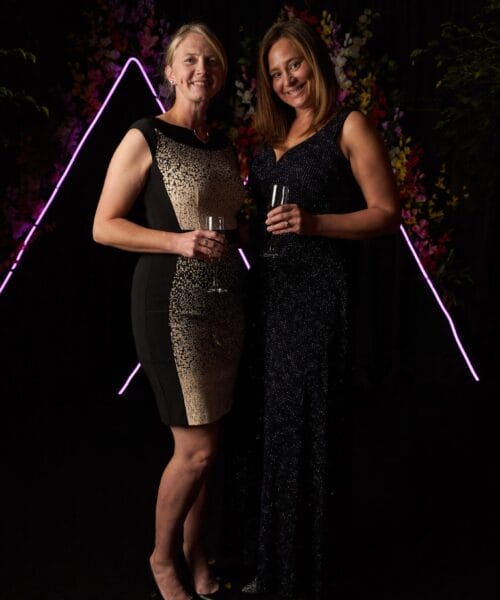 Two smiling women pose together in formal attire at an event. One wears a black and gold dress, while the other dons a black gown. Both hold champagne glasses. The backdrop features colorful flowers and illuminated triangle outlines, creating an elegant atmosphere.