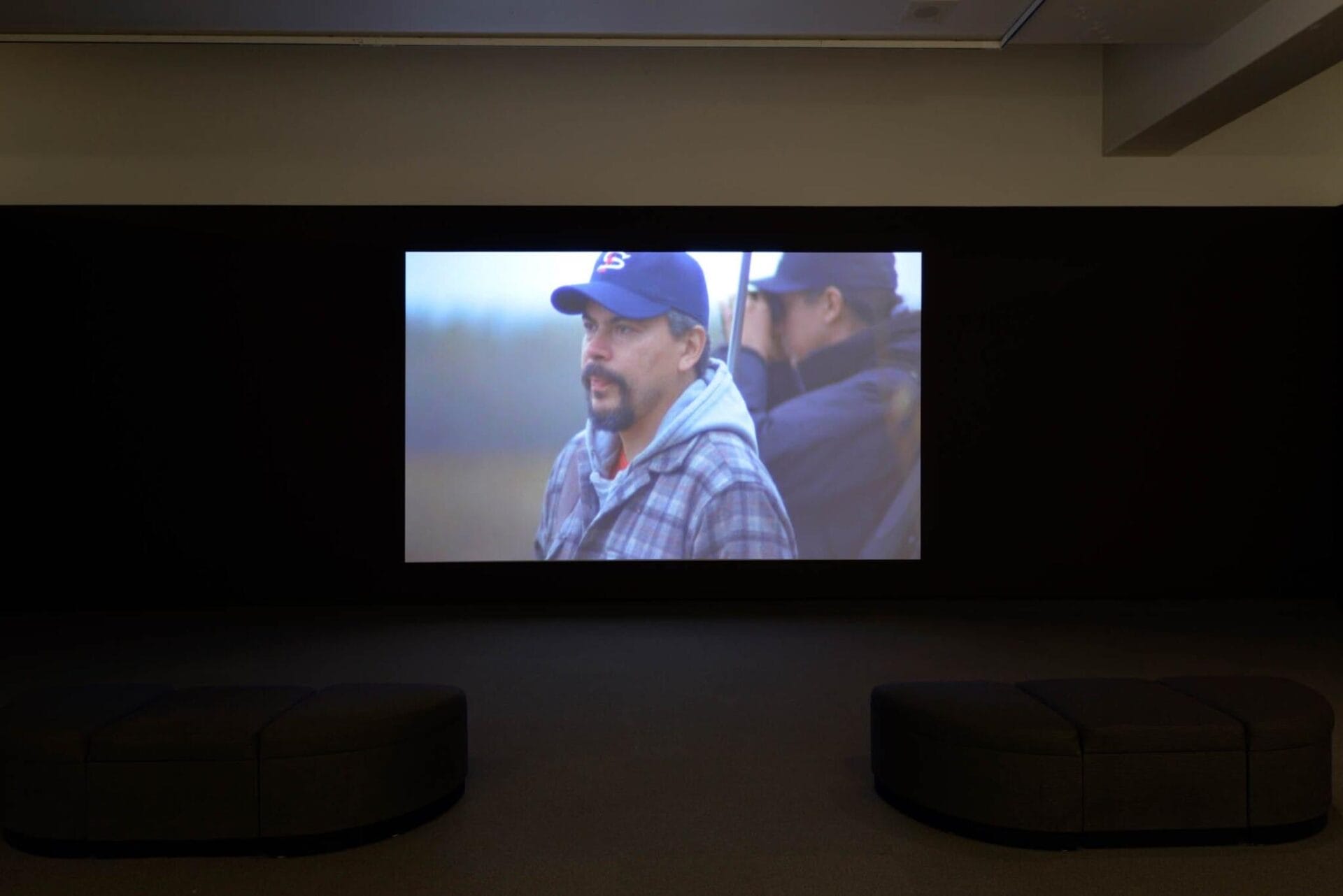 A dimly lit room with a large screen showing a movie scene. The screen displays a close-up of a man with a beard, wearing a blue baseball cap and a plaid shirt, outdoors. The room has two cushioned benches facing the screen.