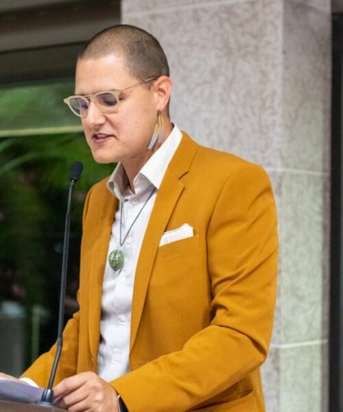 A person with short hair, wearing glasses, a brown blazer, and a white shirt, stands at a podium speaking into a microphone. They have a pendant necklace and an earring. The background shows a stone pillar and greenery.