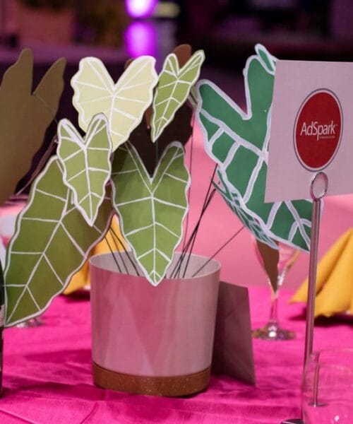A table centerpiece featuring potted green leaf-shaped paper cutouts, a sign with the AdSpark logo, and a wine bottle. The table is covered in a bright pink tablecloth, with wine glasses and yellow napkins around it. The background has a café or event setting.