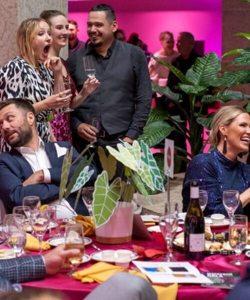 People seated and standing around a table, socializing at a lively event. Some are laughing and engaged in conversations while others are holding drinks. The setting appears elegant with decorative plants and colorful table arrangements.