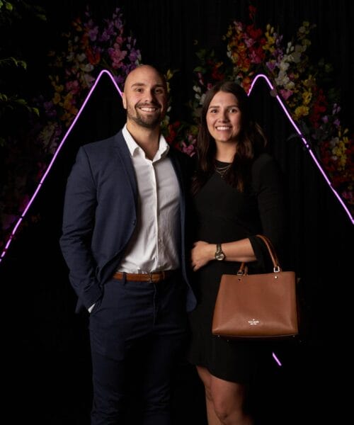 A man and a woman stand together in front of a dark backdrop adorned with neon-like purple triangle shapes and floral decorations. The man is dressed in a navy suit and white shirt, while the woman wears a black dress and carries a brown handbag. Both are smiling.