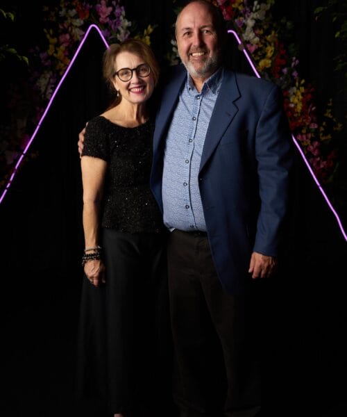 A man and woman stand closely together, smiling at the camera. The woman is wearing glasses, a short-sleeved black top, and a long black skirt. The man is dressed in a blue blazer and dark pants. The backdrop features neon purple triangular lights and colorful flowers.