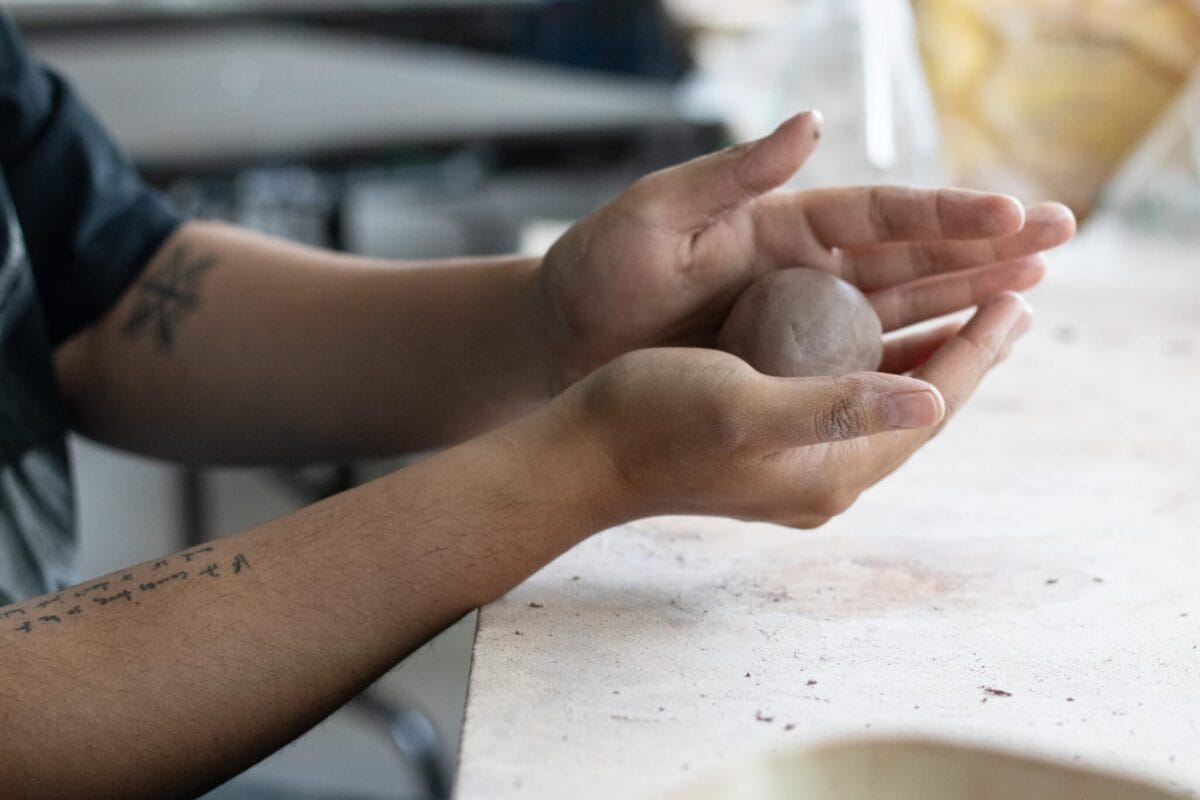 A person with tattoos on their forearms is shaping a clay ball with both hands, creating a Pinch Pot Portrait on a light-colored table. The focus is on the hands and the clay ball, with background details softly blurred.