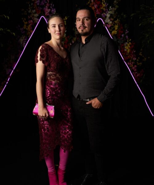 A woman in a burgundy lace dress, pink tights, and pink boots stands next to a man in a gray vest and black shirt with black pants. They are posing in front of a dark background with colorful flowers and neon lines shaped like mountains in the backdrop.
