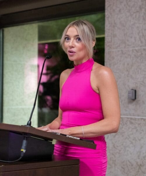 A woman in a bright pink sleeveless dress stands at a podium, speaking into a microphone. She has blonde hair pulled back and is looking ahead, with her hands resting on the sides of the podium. The background features a textured wall and a window.
