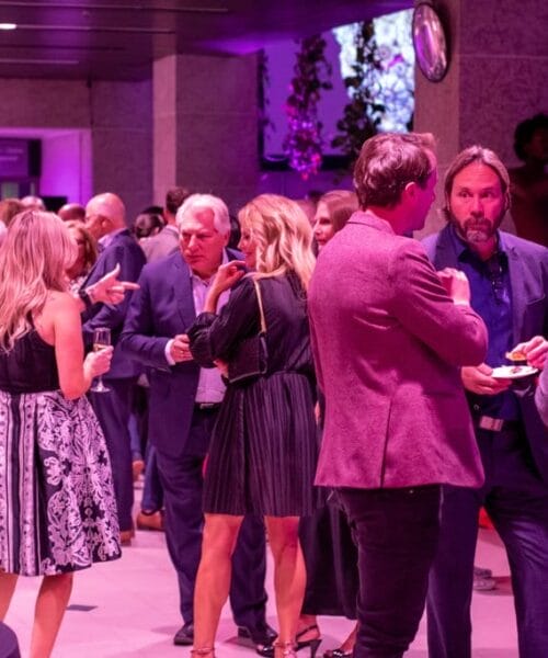 A group of people in formal attire socializing at an indoor event with purple lighting. Some are holding drinks and engaging in conversations, while others are eating from plates. Party decorations and a few TV screens in the background add to the ambiance.