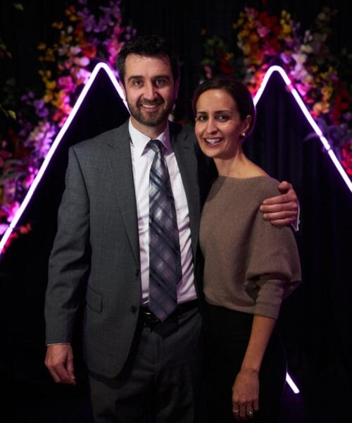A man in a suit and tie stands with a woman in a beige top, arm around her shoulder. They are smiling in front of a colorful floral backdrop illuminated by triangular neon lights. The setting appears formal and celebratory.