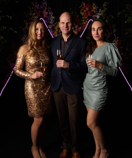 Three people dressed in formal attire pose against a floral backdrop with pink neon lights. The two women wear elegant dresses, one in gold and the other in silver, while the man in the center wears a dark blazer. They all hold champagne glasses and smile at the camera.