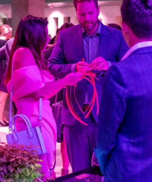 A group of people stand around a table at a social event illuminated by purple light. A man in a suit inspects some items, while a woman in a pink outfit with a blue handbag looks on. Other attendees are visible in the background, and a table with objects is in the foreground.