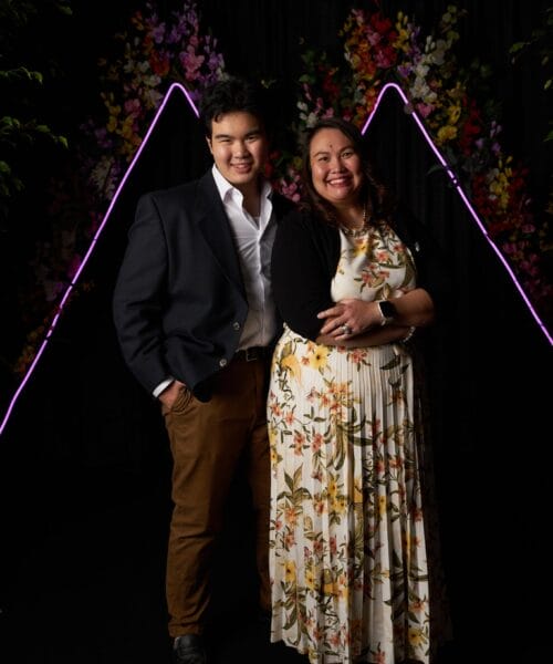 A man and a woman stand side by side, smiling. The man wears a dark blazer, white shirt, and brown pants. The woman wears a floral dress and black cardigan. They stand in front of a black backdrop with colorful floral decorations and purple neon lights forming triangular shapes.