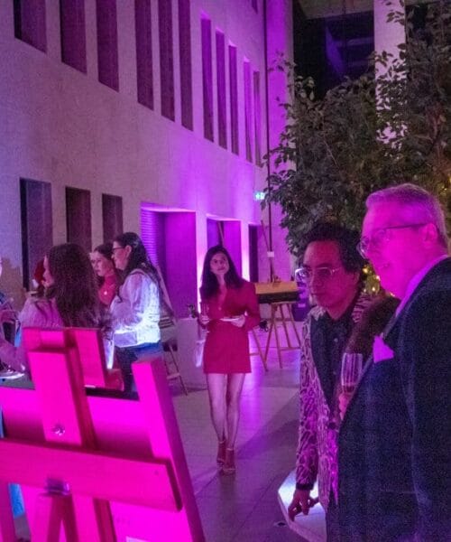 A group of people are gathered at an art gallery with soft purple lighting. They are viewing various paintings displayed on easels. Two men in suits are in the foreground, engaged in conversation while others admire the artwork. A large digital screen is visible on the wall.